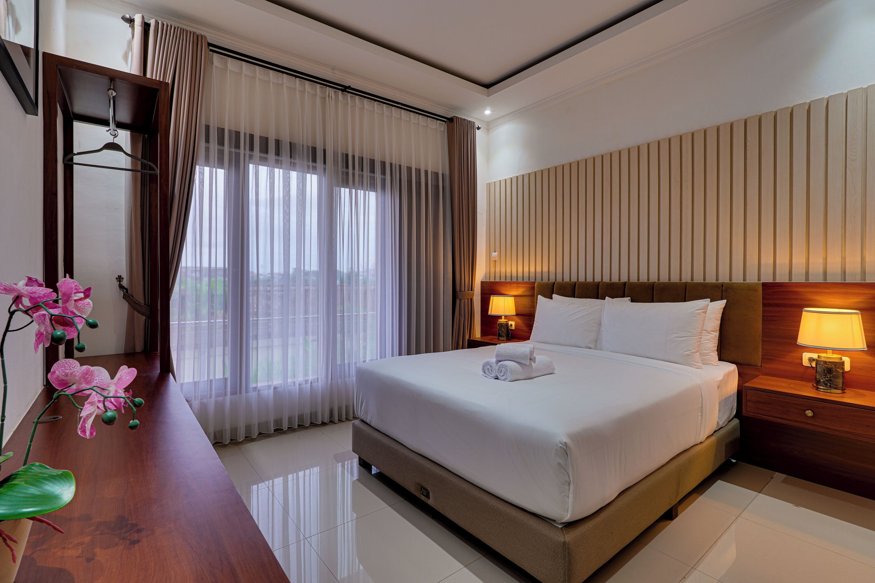 Une chambre d’hôtel avec un grand lit, un bureau en bois, une lampe et une vue sur l’extérieur à travers des rideaux transparents.