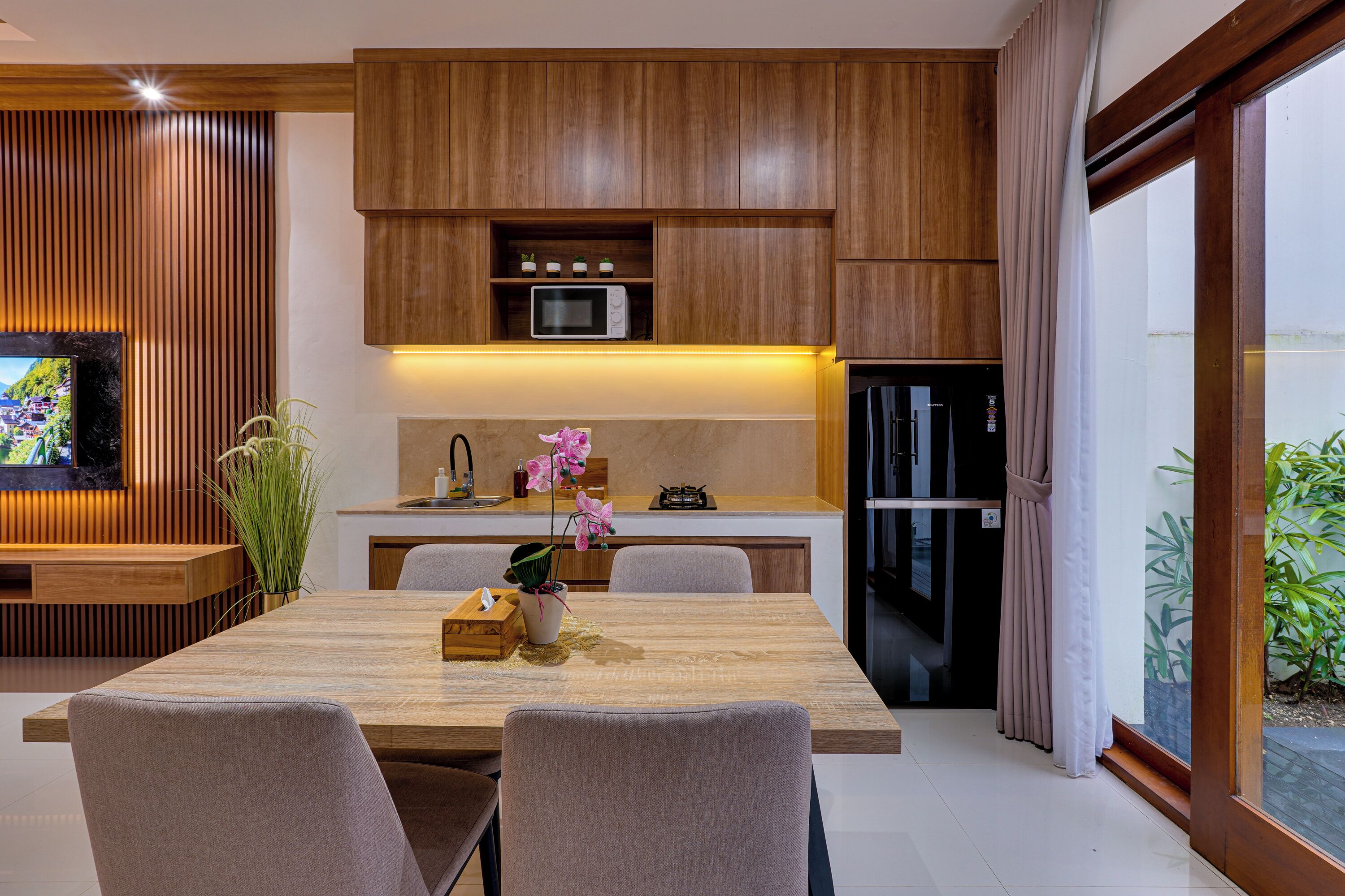 A modern kitchen with wooden cabinets, a microwave, and a refrigerator. There is a dining table with chairs and a vase of flowers.