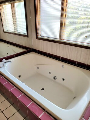 Une salle de bain avec une grande baignoire, un miroir et deux fenêtres équipées de stores.