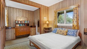 A room with wooden panel walls, a bed with a white mattress and floral pillows, a wooden desk with books, and a window with curtains. - Elanora is an original cottage (Bendalong)