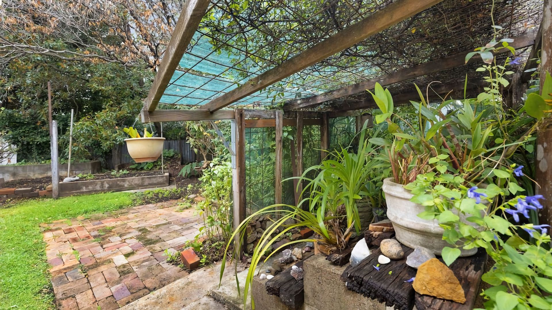 Un jardí amb un camí de maó, planters penjants i una estructura de pergola de fusta.