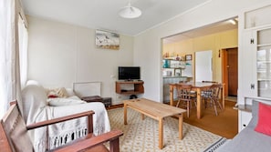 A living room with a sofa, TV, wooden table, and a dining area with chairs. - Elanora is an original cottage (Bendalong)
