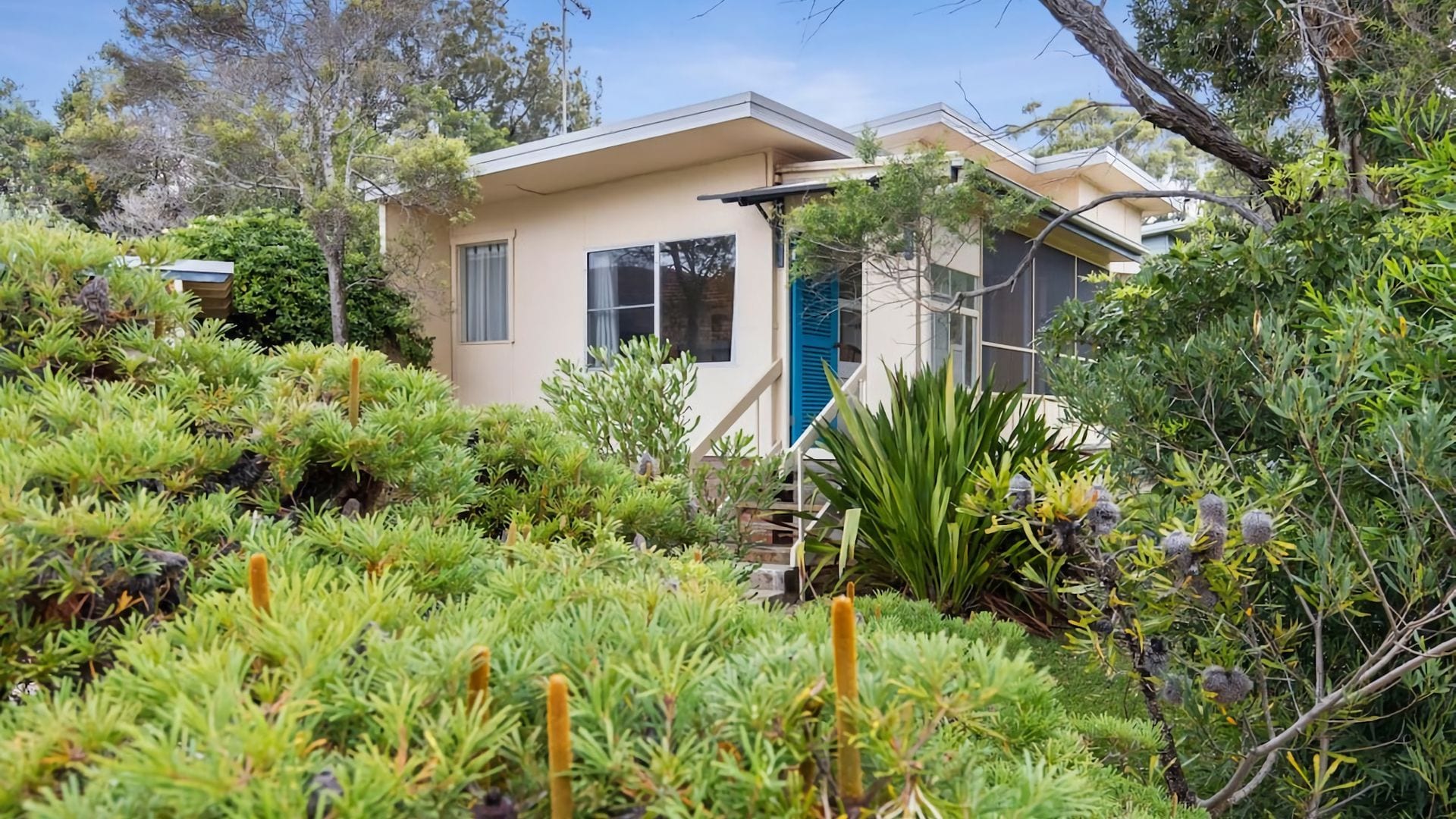 Une maison moderne à l’extérieur beige, entourée d’une végétation luxuriante et d’arbres.