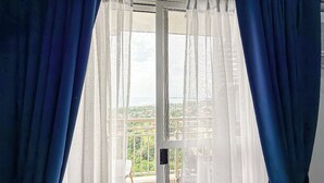 A window with sheer white curtains and solid blue curtains, offering a view of a balcony with greenery and buildings in the distance. - Verdon Parc Davao City Condo - Perfect Staycation Near SM Ecoland (Davao City)