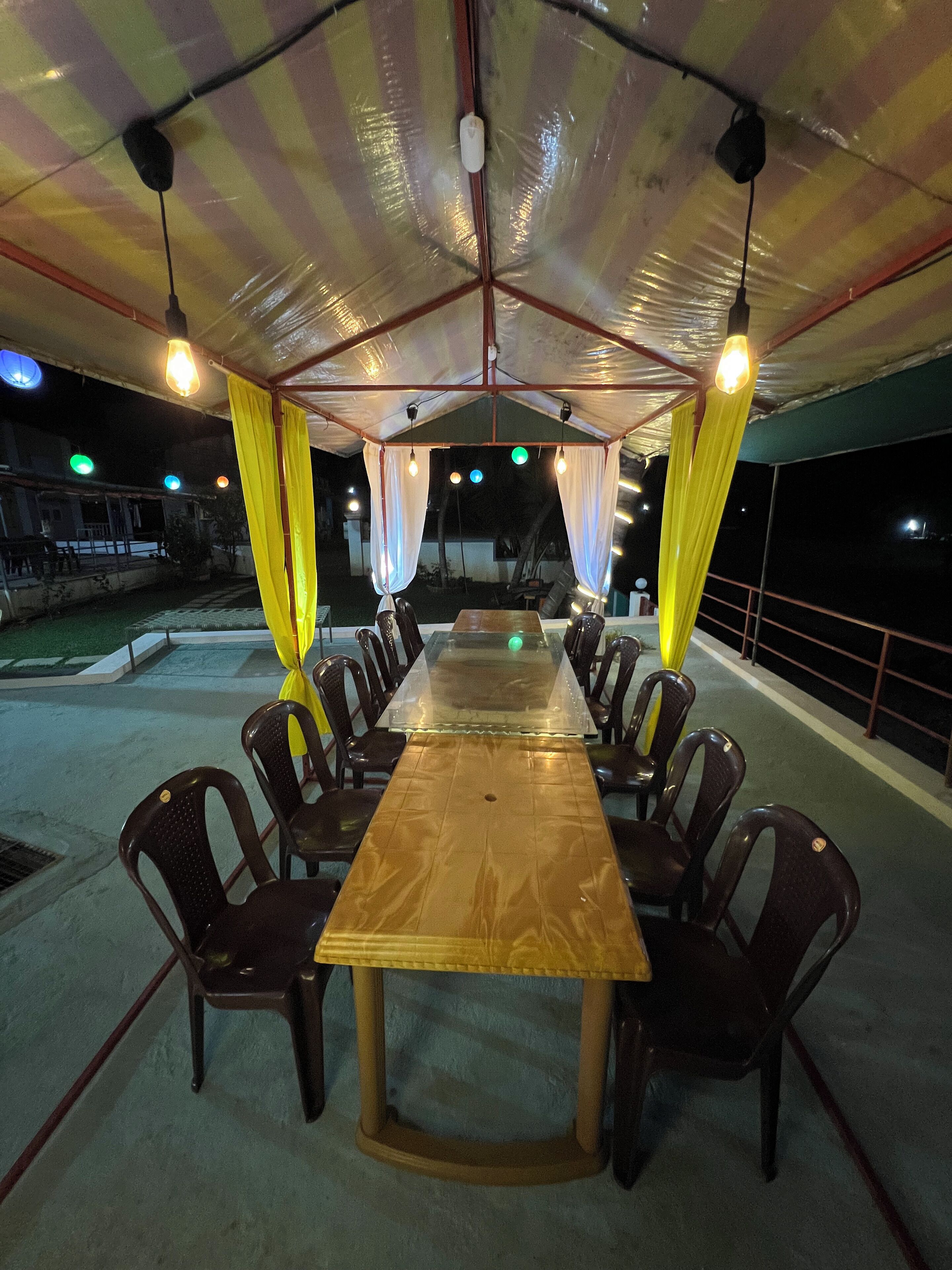 A covered outdoor dining area with a long wooden table and chairs, under a striped canopy with hanging lights.