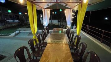 A covered outdoor dining area with a long wooden table and chairs, under a striped canopy with hanging lights.