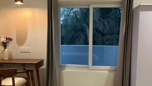 A bedroom with a bed, a desk with a chair, a window with curtains, and a hanging light fixture. - Noi Homes (East Jorhat)