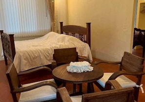 A room with a bed, a round wooden table with chairs, and a window with curtains.