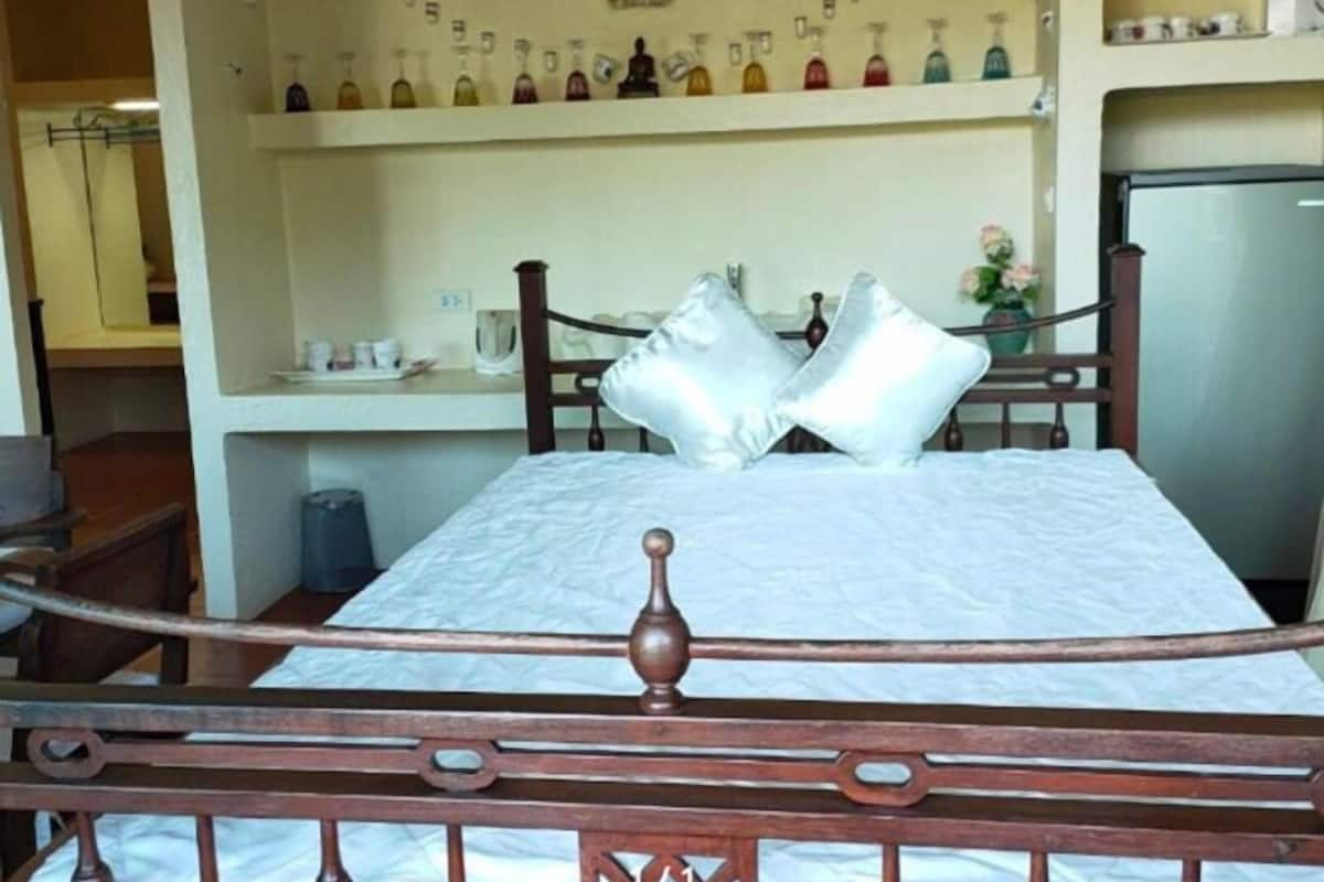 A bedroom with a wooden bed frame, a white mattress, and a shelf above the bed holding various bottles.