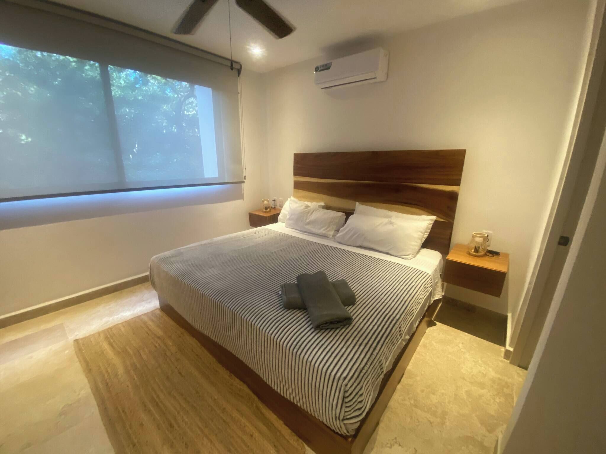 A modern bedroom with a large bed, wooden headboard, bedside tables, and a window with a view of trees.