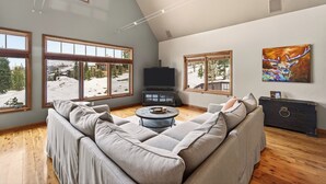 A spacious living room with a large gray sectional sofa, a round coffee table, and a flat-screen TV. There are large windows offering a view of a snowy landscape. - Winter Park Getaway w/ Fireplace, Game Room & Easy Resort Bus | Elk Court Lodge (Winter Park)