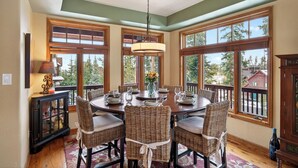 A dining area with a round table set for six, surrounded by wicker chairs, large windows with wooden frames, and a hanging light fixture. - Winter Park Getaway w/ Fireplace, Game Room & Easy Resort Bus | Elk Court Lodge (Winter Park)