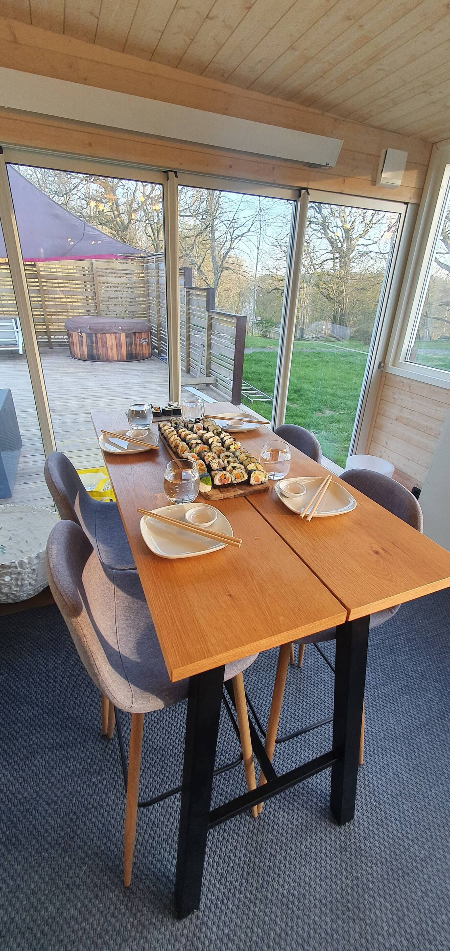 A dining area with a wooden table set for two, a large window with a view of the outdoors, and a wooden deck visible through the window.