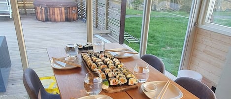A dining area with a wooden table set for two, a large window with a view of the outdoors, and a wooden deck visible through the window.