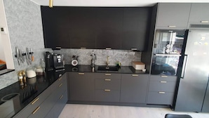 Cozinha moderna com armários escuros, bancada preta e eletrodomésticos de aço inoxidável, incluindo uma geladeira e uma cafeteira.