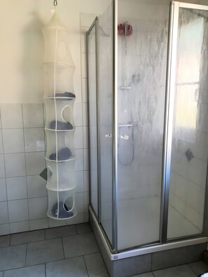 Ett badrum med en dusch som är avskärmad med glasväggar, vita kakelväggar och ett vitt vägghängt hyllsystem.