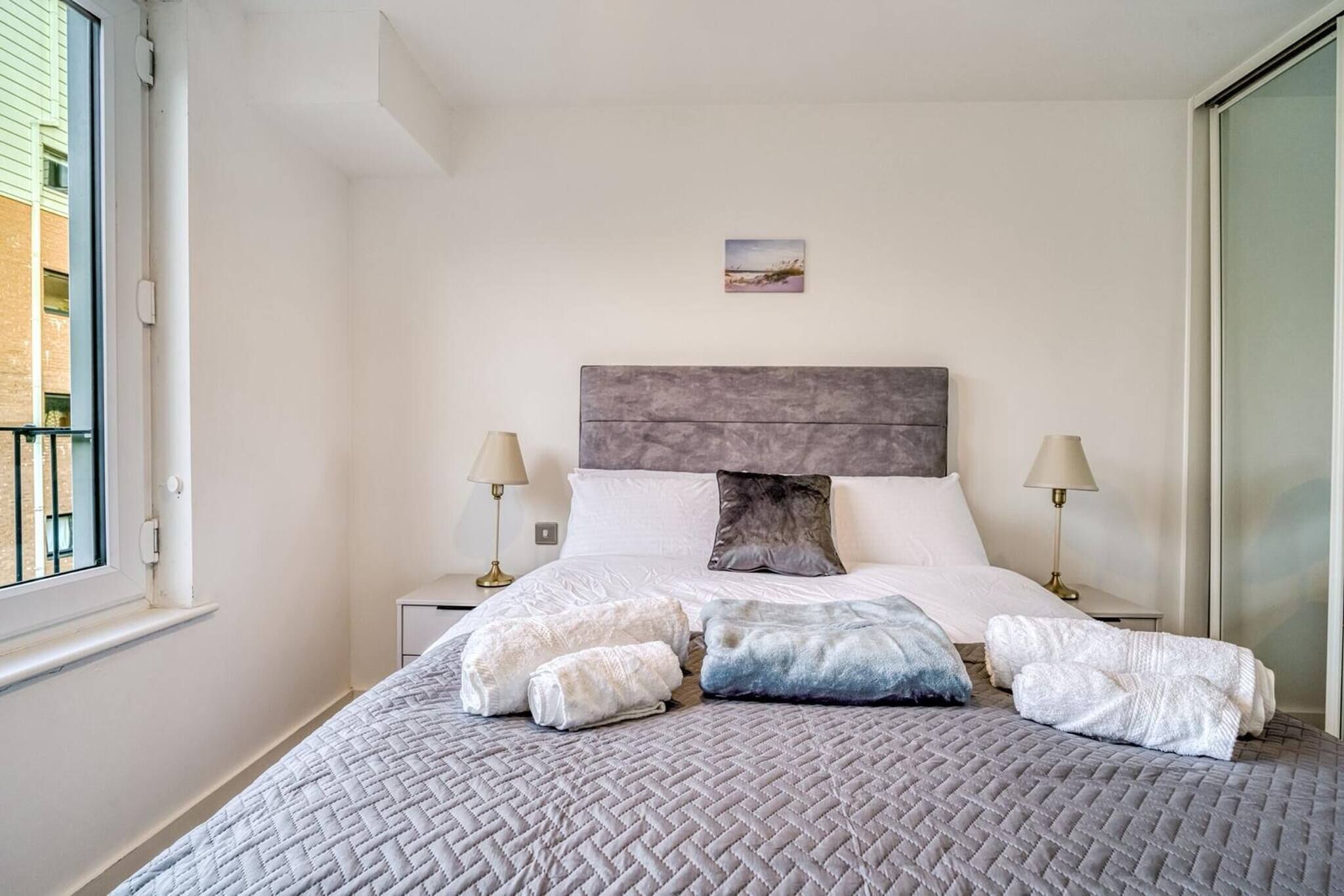 Un lit bien fait, recouvert d’une courtepointe grise matelassée, avec deux lampes de chevet et un petit tableau accroché au mur.