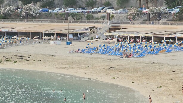 Una playa con hileras de tumbonas azules y blancas, un cartel que indica servicios como duchas e inodoros, y una vista despejada del mar.