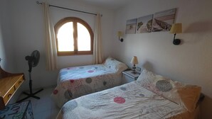 Un dormitorio con dos camas, un escritorio, un ventilador de pie, una ventana con cortinas y lámparas fijadas a la pared.