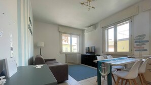A modern, well-lit room with a dining table, chairs, a sofa, a TV, and a large window providing natural light. - Charming Canal-side Apartment in Cesenatico, Italy (Cesenatico)