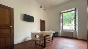 Ein Zimmer mit Holztisch und -stühlen, einem flachbildfernseher an der Wand, einem Fenster mit Blick ins Grüne und einer Heizung unter dem Fenster.