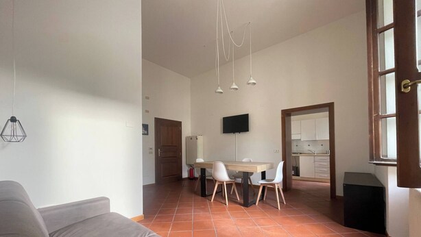 Ein Wohnzimmer mit einer Couch, ein Essbereich mit Tisch und Stühlen, eine durchgehende Küche hinter einer offenen Tür und Pendelleuchten an der Decke.
