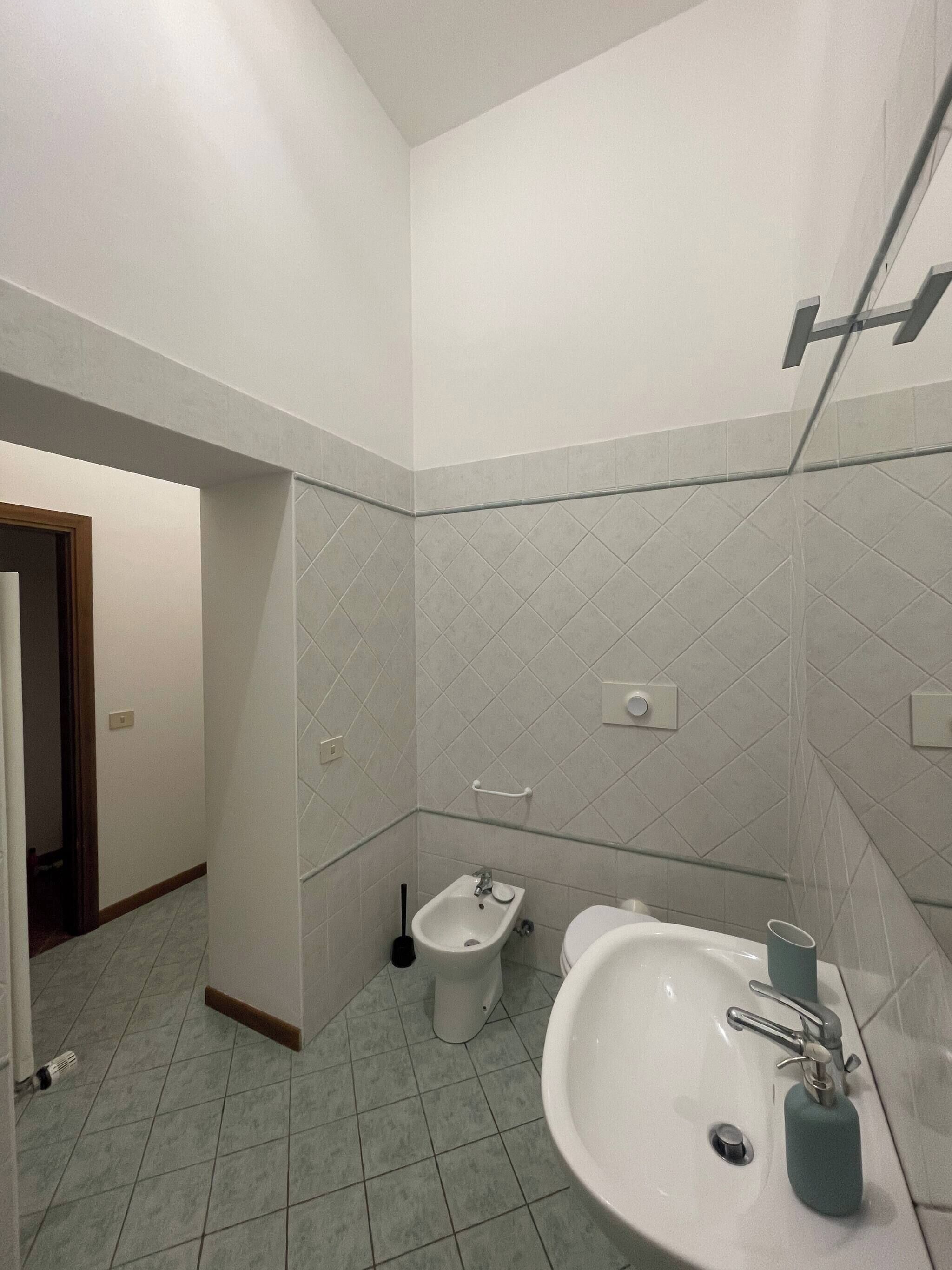 Een badkamer met een wastafel, toilet en bidet. De wanden zijn betegeld en er is een deur naar een andere kamer.