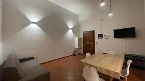 Ein modernes Zimmer mit Esstisch aus Holz, weißen Stühlen, einer Couch, einem Flachbildfernseher und Pendelleuchten.
