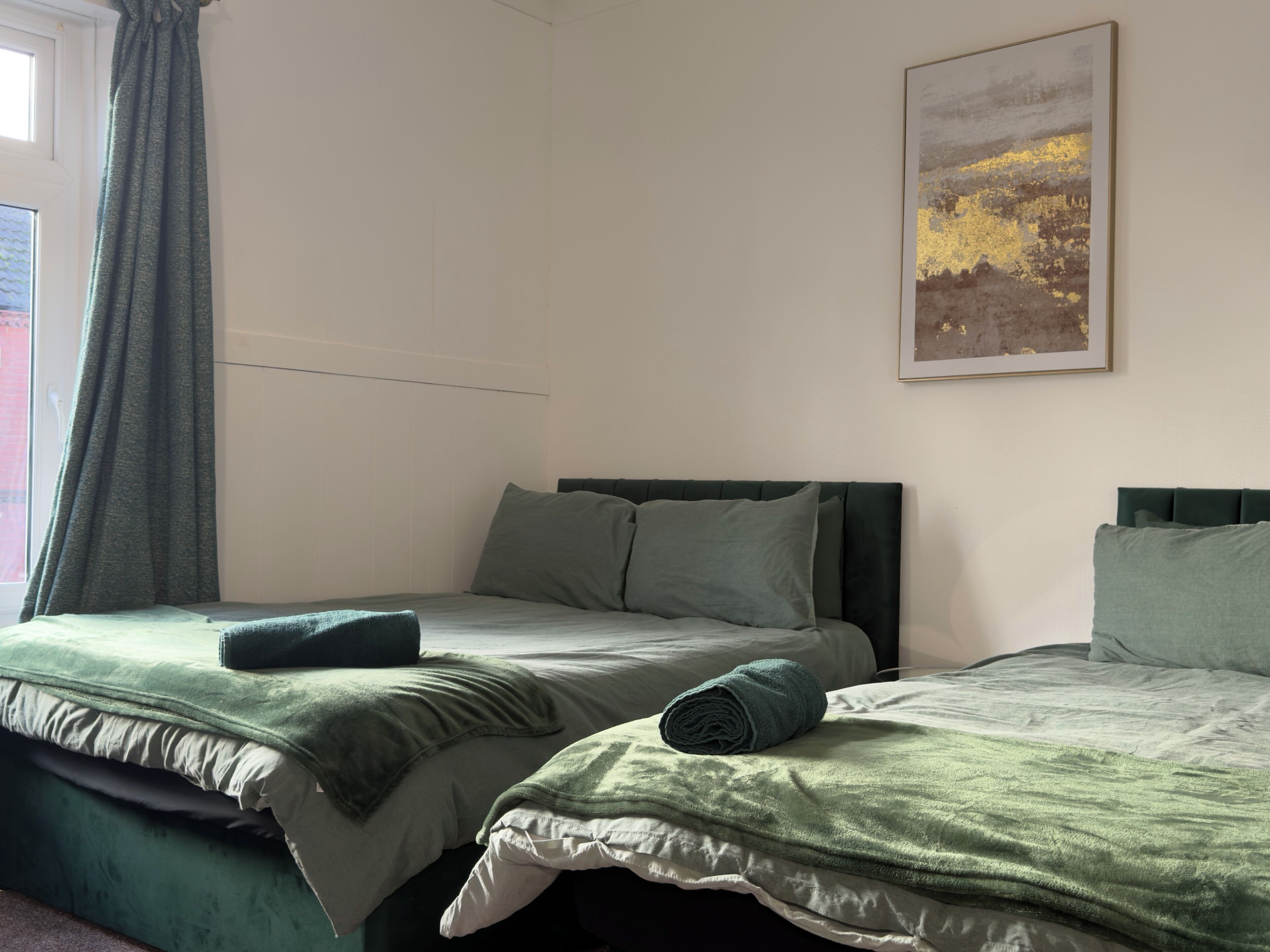 Habitación de hotel con dos camas, cada una con ropa de cama y almohadas verdes, una ventana con cortinas y un cuadro en la pared.