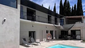 Un edificio moderno con piscina, mobiliario exterior y altos árboles al fondo.
