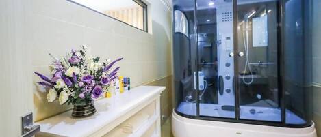 Un baño moderno con ducha envuelta en vidrio, tocador blanco con un jarrón de flores y un espejo grande sobre el tocador.