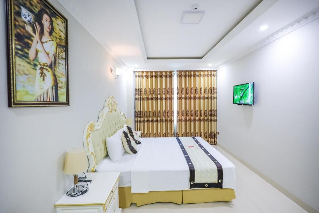 A hotel room with a large bed, a bedside table with a lamp, a television on the wall, and a framed picture above the bed.