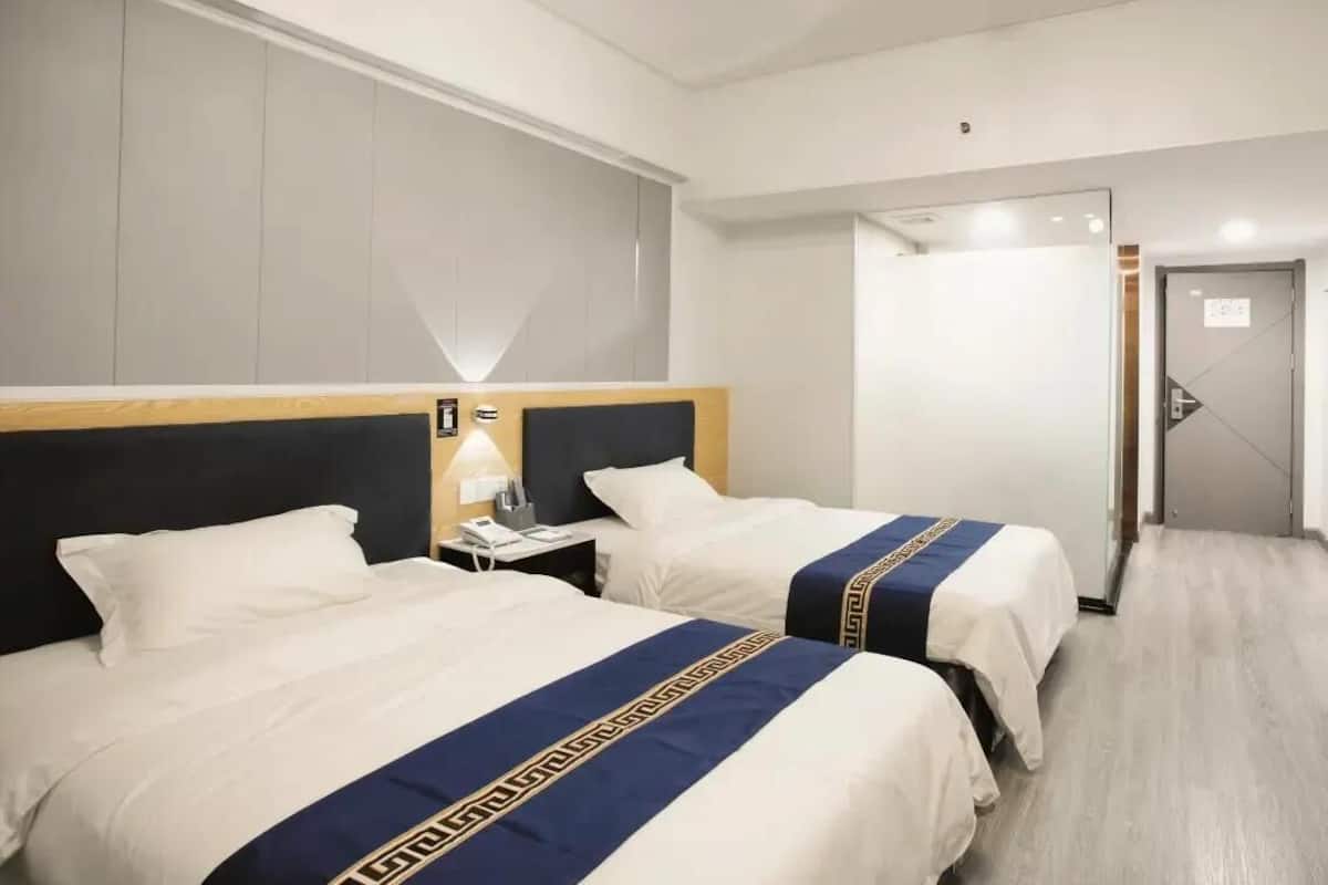 A hotel room with two beds, a nightstand with a lamp, a wooden headboard, and a door leading to a bathroom.
