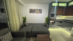 Ein modernes Wohnzimmer mit einem grauen Ecksofa, einem kleinen Holztisch und einer Küchenzeile mit Edelstahlgeräten.