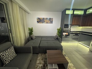 Ein modernes Wohnzimmer mit einem grauen Ecksofa, einem kleinen Holztisch und einer Küchenzeile mit Edelstahlgeräten.
