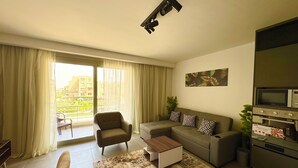 Ein modernes Wohnzimmer mit einer Couch, einem Sessel und einem Couchtisch. Es gibt ein großes Fenster mit Vorhängen, eine Deckenleuchte und einen wandmontierten Backofen.