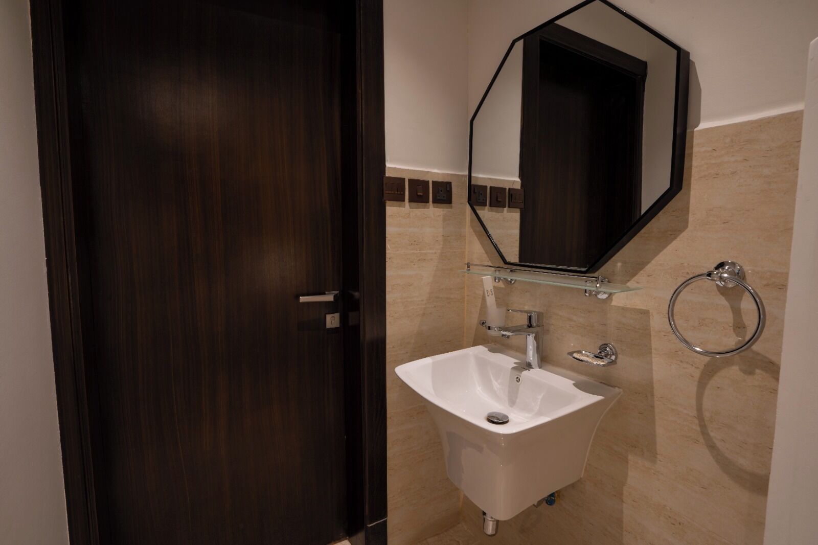 A bathroom with a dark wooden door, a white sink, a glass shelf above the sink, a mirror, and a towel ring.