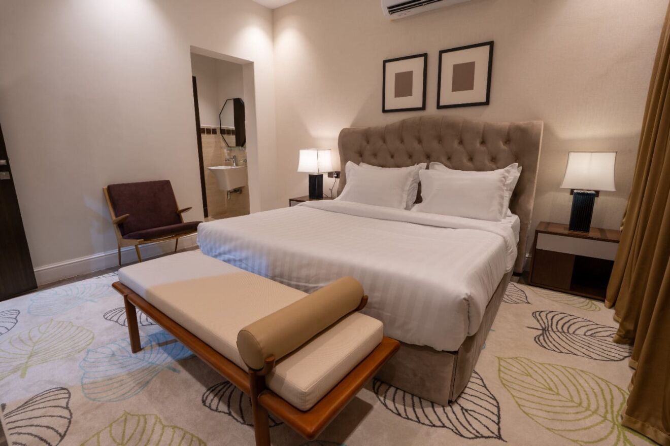 Hotellihuone, jossa on suuri sänky, yöpöytä lampun kanssa, tuoli ja peili.