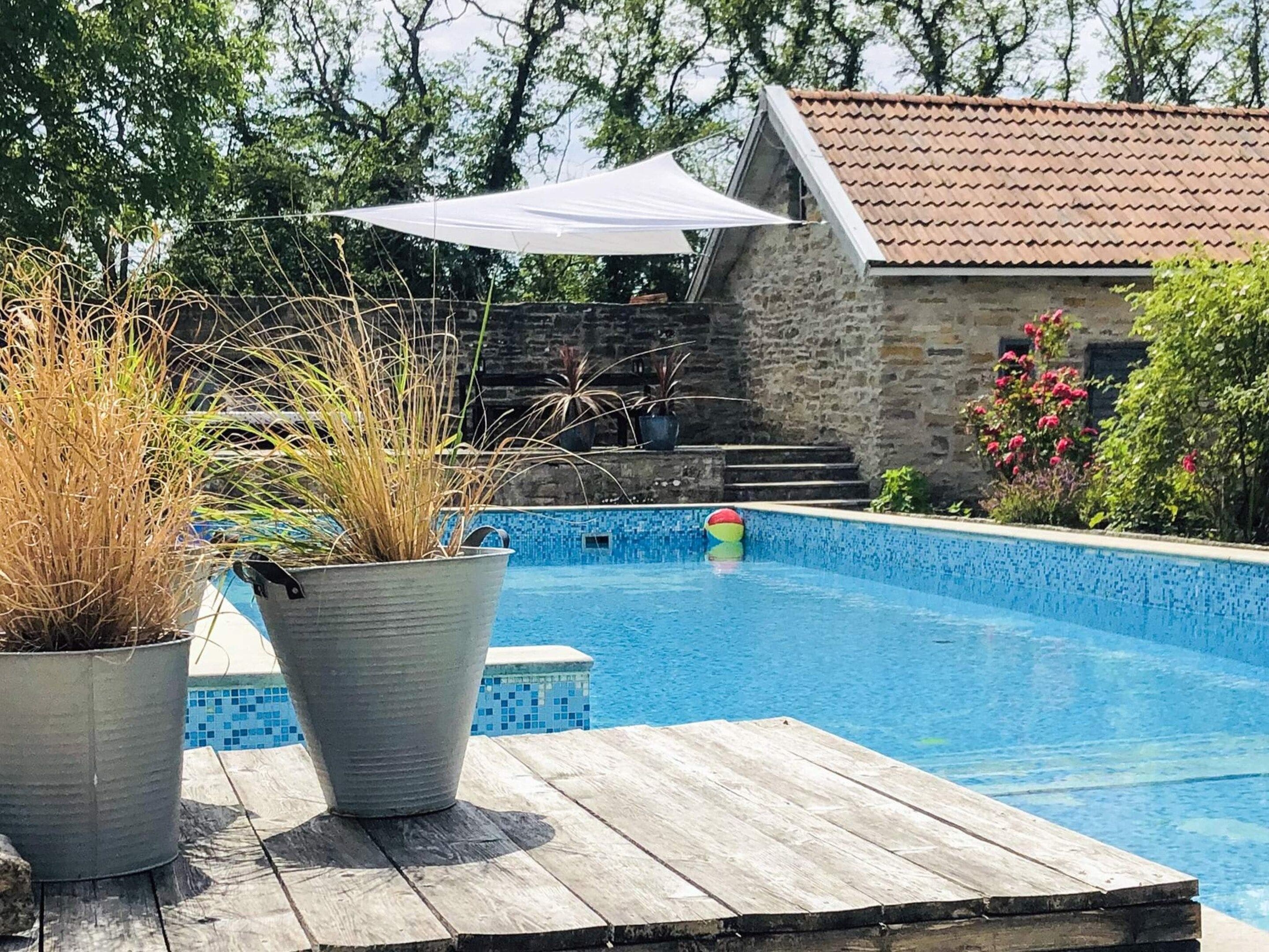 Una piscina amb un pont de fusta davant, envoltada de plantes en testos i una paret de pedra.