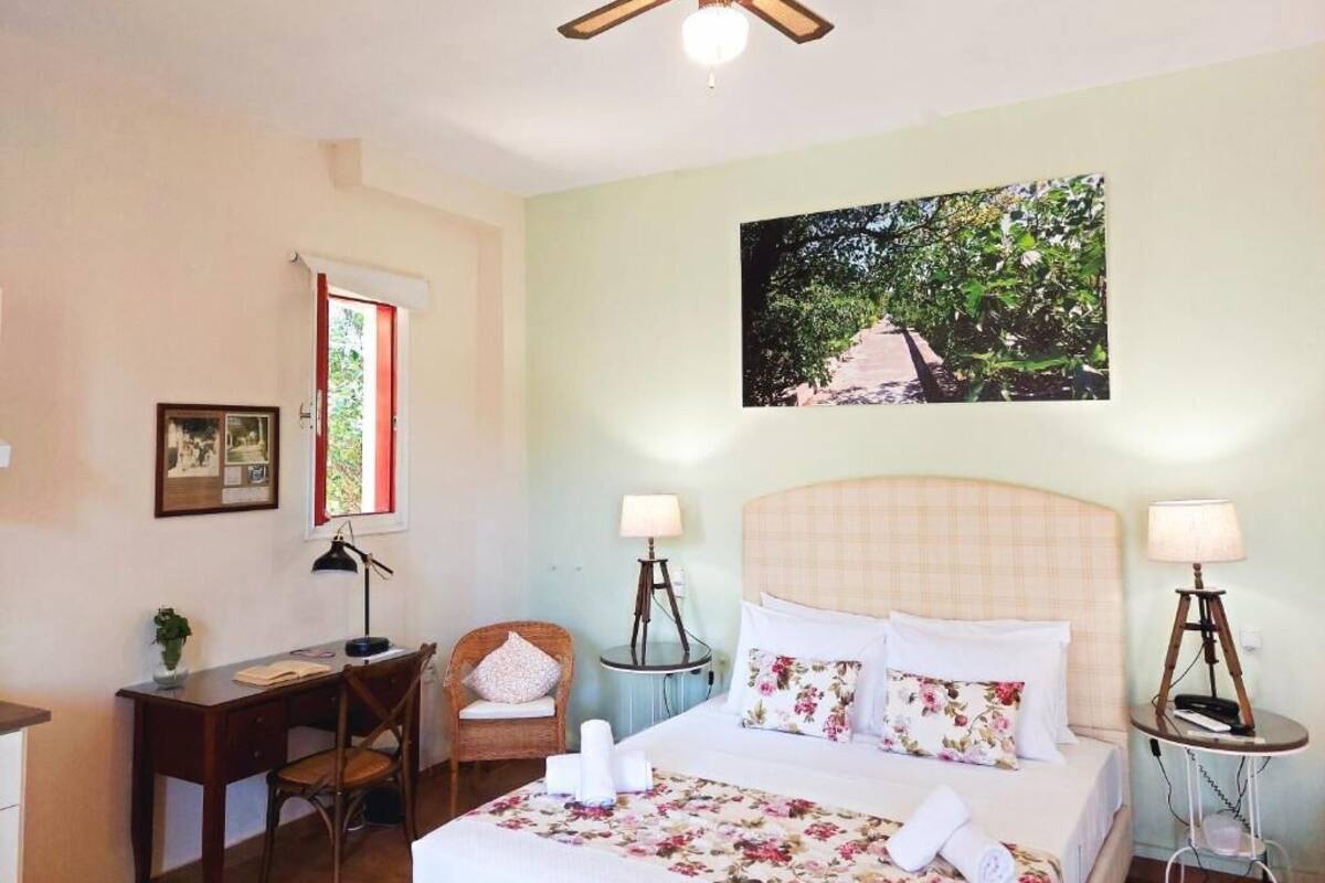 A bedroom with a bed, two bedside tables with lamps, a desk, a chair, a window with red curtains, and a ceiling fan.