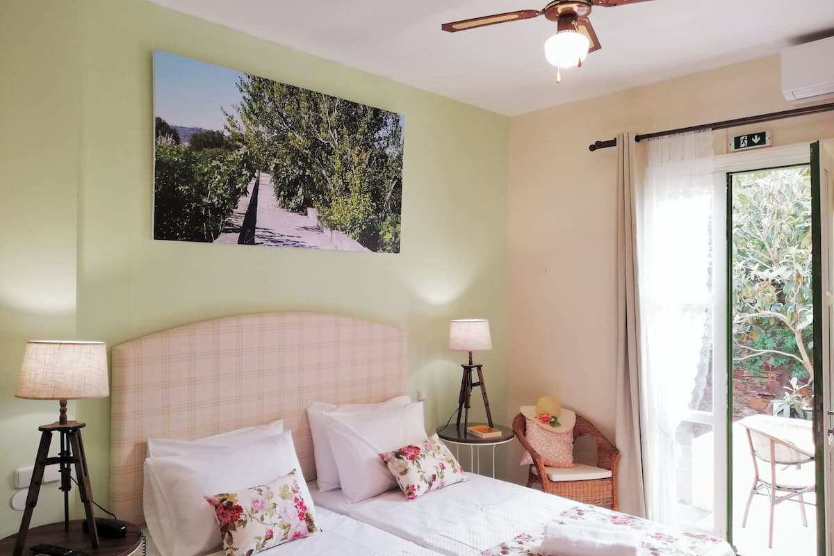 A hotel room with two beds, a ceiling fan, a painting of trees, a chair, and a view of the outdoors.