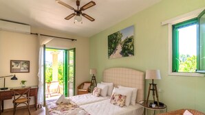 A bedroom with two beds, a desk, a chair, a ceiling fan, and a window with a view of greenery. - BELLA CISTERNA (Chios)