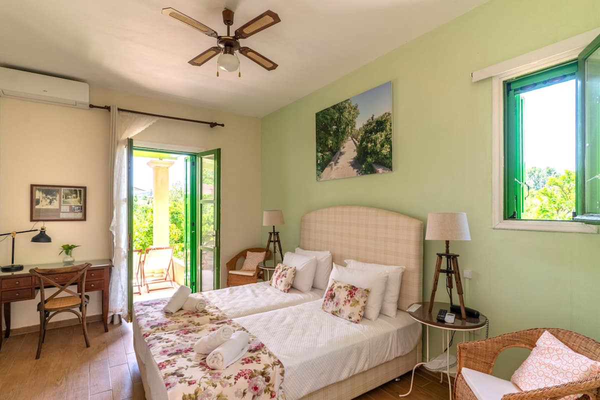 A bedroom with two beds, a desk, a chair, a ceiling fan, and a window with a view of greenery.
