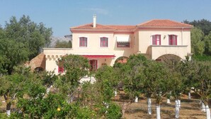 A two-story house with a red-tiled roof, a balcony, and a garden with trees and shrubs. - BELLA CISTERNA (Chios)