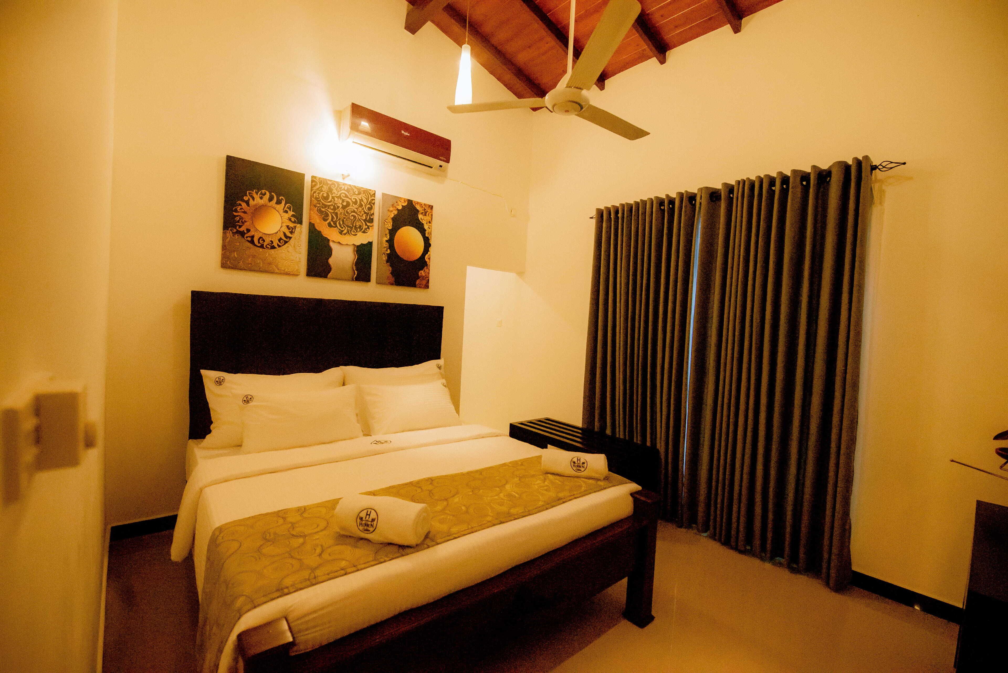 A hotel room with a bed, a ceiling fan, a window with curtains, and artwork on the wall.