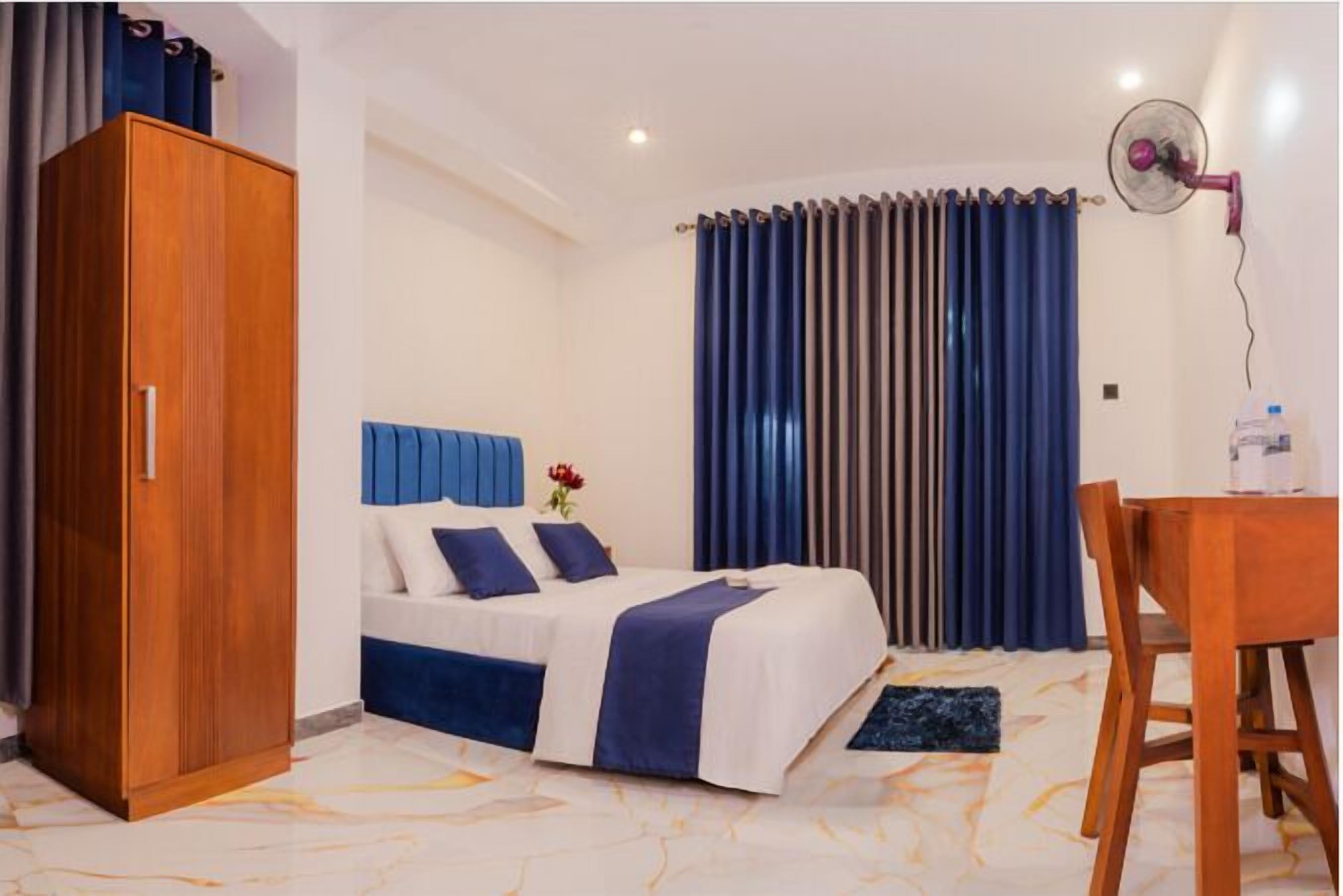 A hotel room with a bed, a wooden wardrobe, a dining table, and a fan.