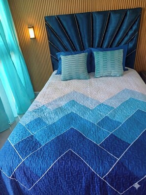 Una cama con colcha a cuadros azules y blancos, dos almohadas a juego y un cabecero tapizado oscuro.