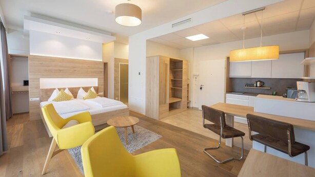 A modern hotel room with a bed, yellow chairs, a small table, a kitchen area, and a dining table. - Comfort Studio With King-size Bed for 2 People 24-7 Check-in (Graz)