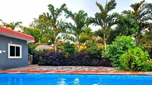 A swimming pool with a tiled deck, surrounded by lush greenery and palm trees. - Arika Farm House (Indore)