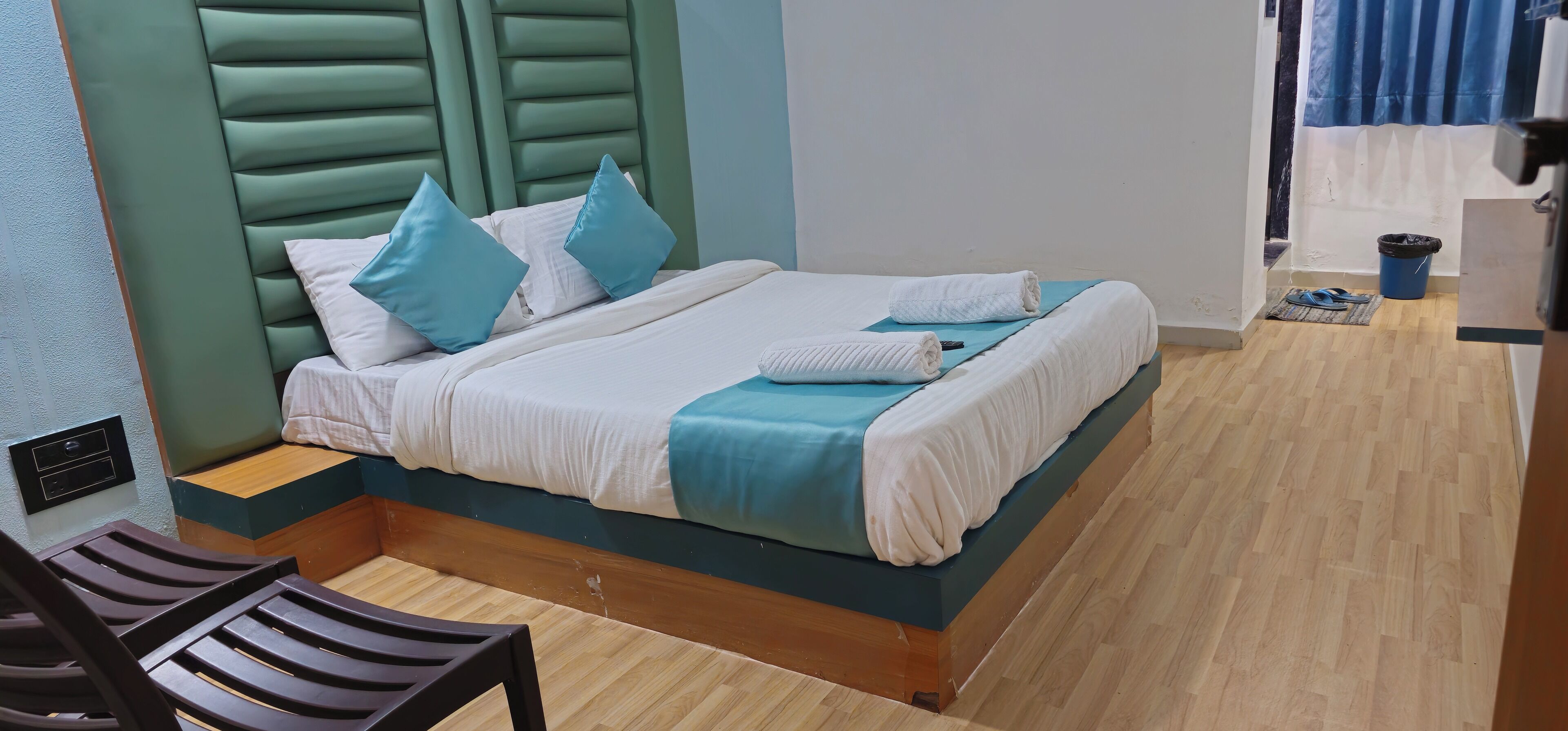 Cama bem arrumada com travesseiros e cobertor, chão de madeira, uma cadeira e uma pequena lixeira.